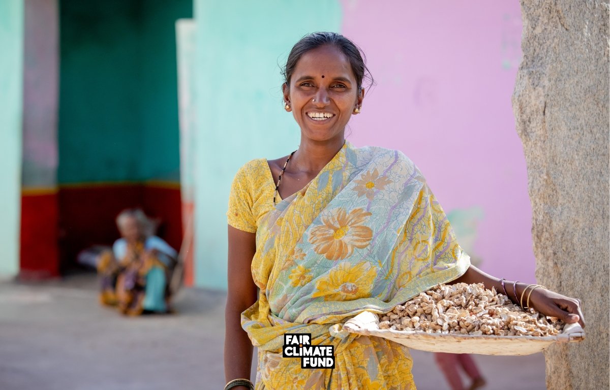 Our approach to social impact A woman in a yellow sari smiles, holding a woven tray with nuts. A person sits in the background against a pastel-colored wall. "Fair Climate Fund" is printed on the image. Designed by Circular Design, a not-for-profit web design agency.