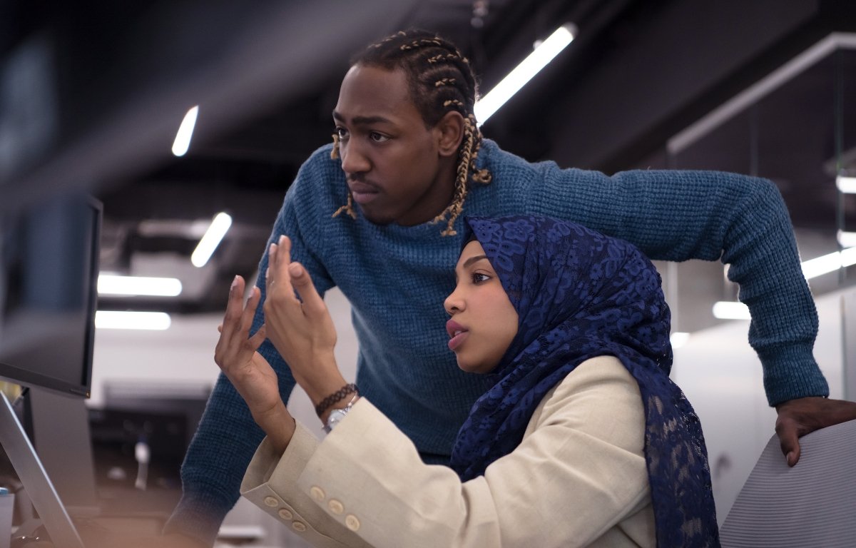 Project Manager Image Two people in an office setting look intently at a computer screen. The woman, wearing a hijab, gestures with her hand, while the man leans forward with his hand on his hip. Designed by Circular Design, a not-for-profit web design agency.