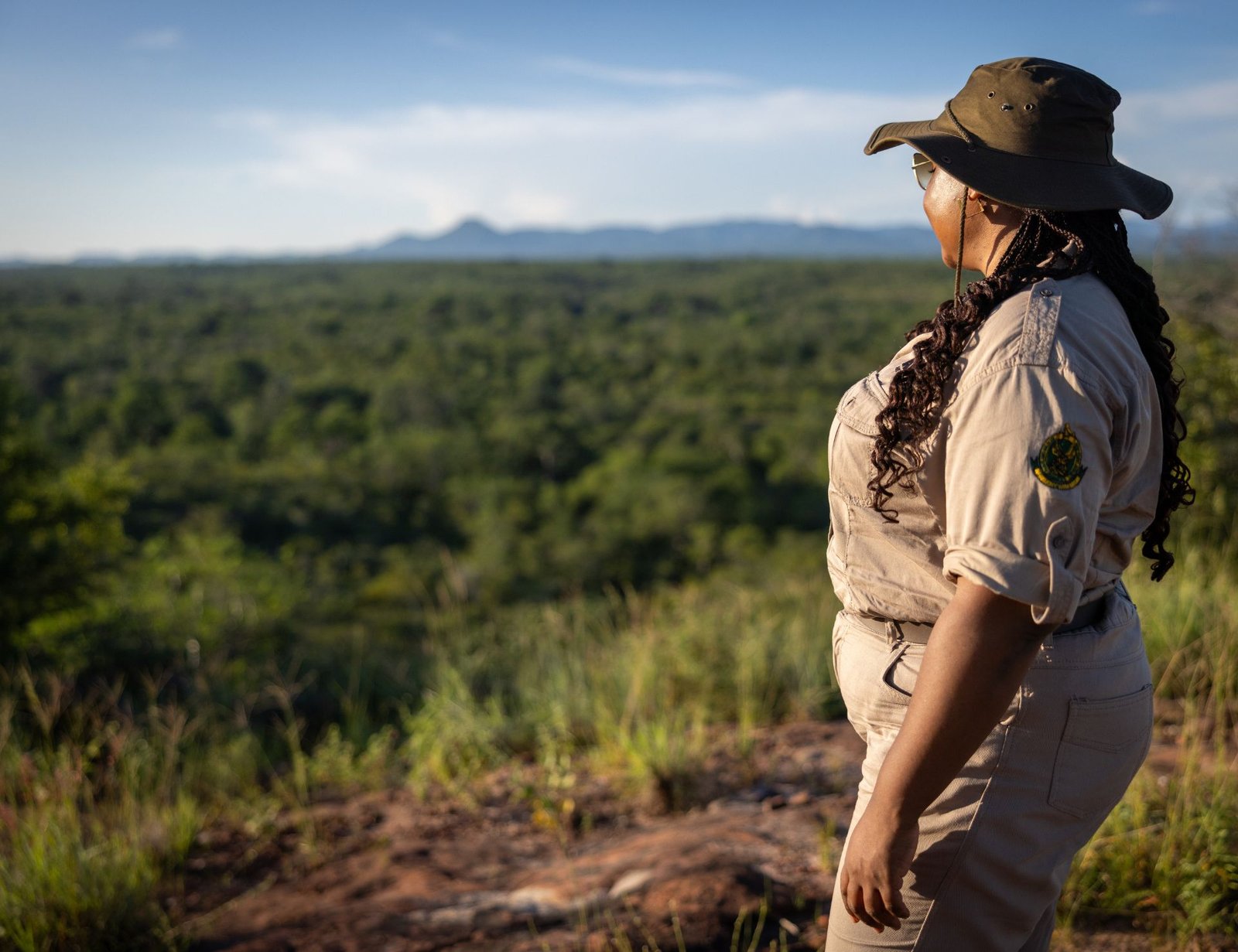 AWE for Nature A person in a safari uniform and hat stands on a rocky hill, looking out over a vast, green landscape under a clear blue sky. Designed by Circular Design, a not-for-profit web design agency.