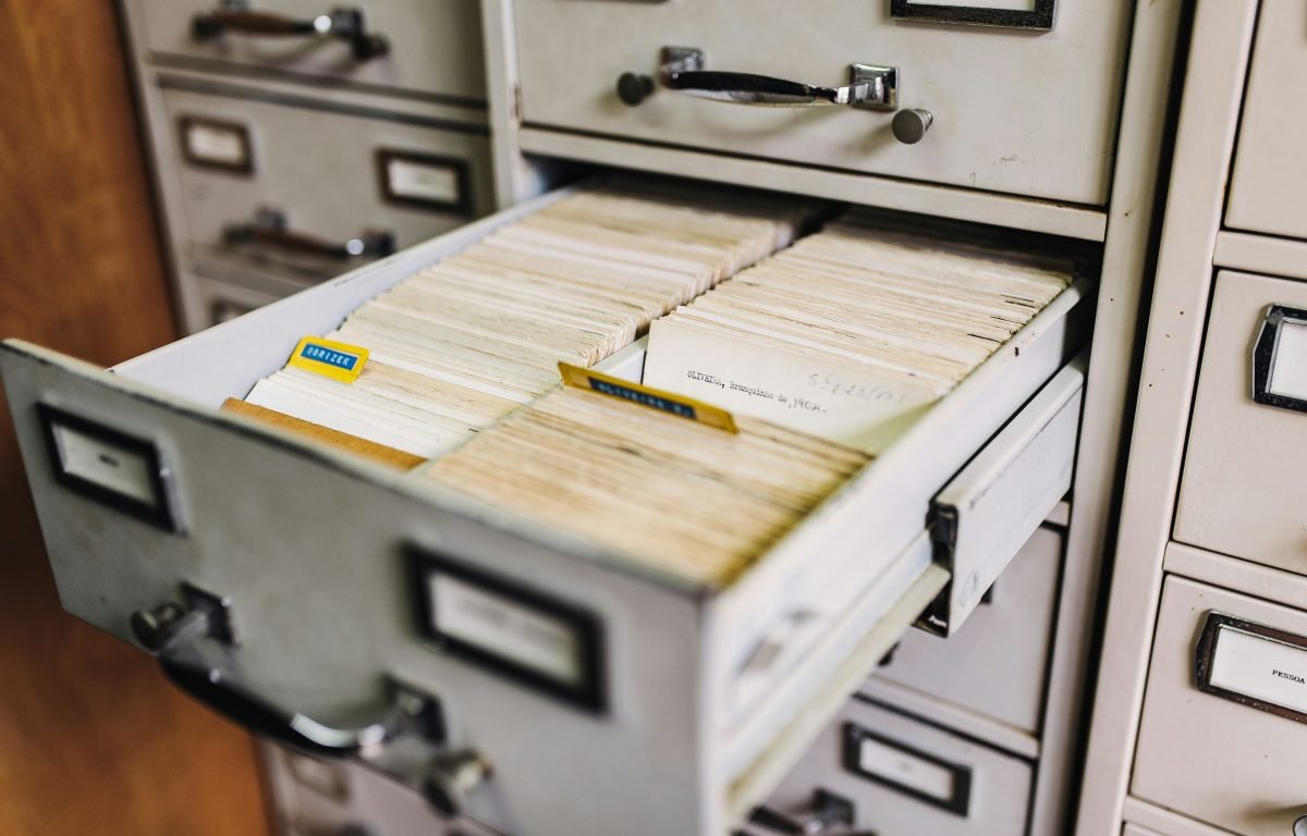 content-management-system-scaled_ An open metal filing cabinet drawer filled with organized index cards, some labeled with colored tabs, in a room with similar cabinets.